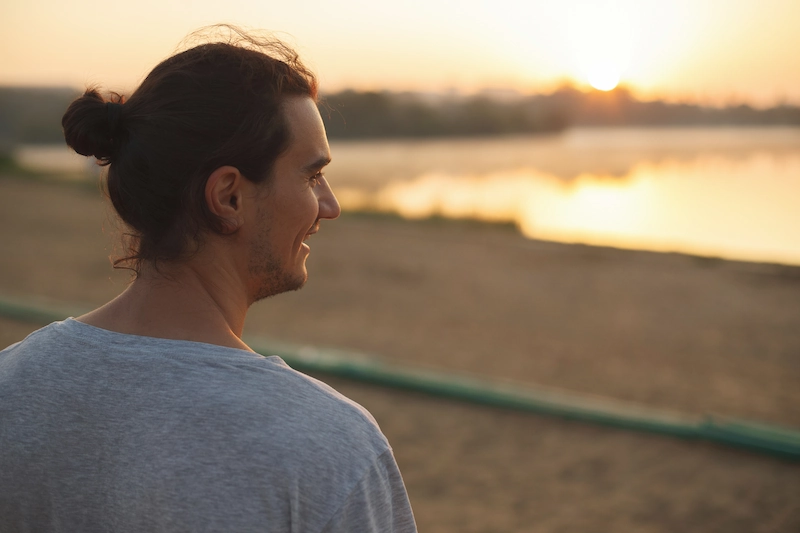 Knappe man met knotje lacht terwijl hij in het park naar de zonsondergang bij het water kijkt.
