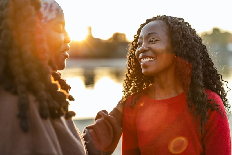 Twee vrouwen praten en lachen samen bij een ondergaande zon aan het water.