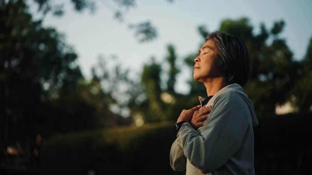 Vrouw staat in de natuur met gesloten ogen en handen op haar borst.