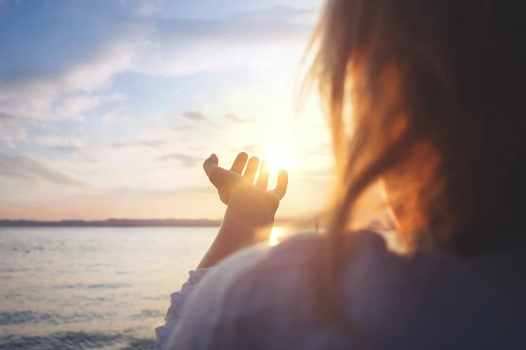 Vrouw reikt over haar schouder naar de ondergaande zon boven een kalme zee.